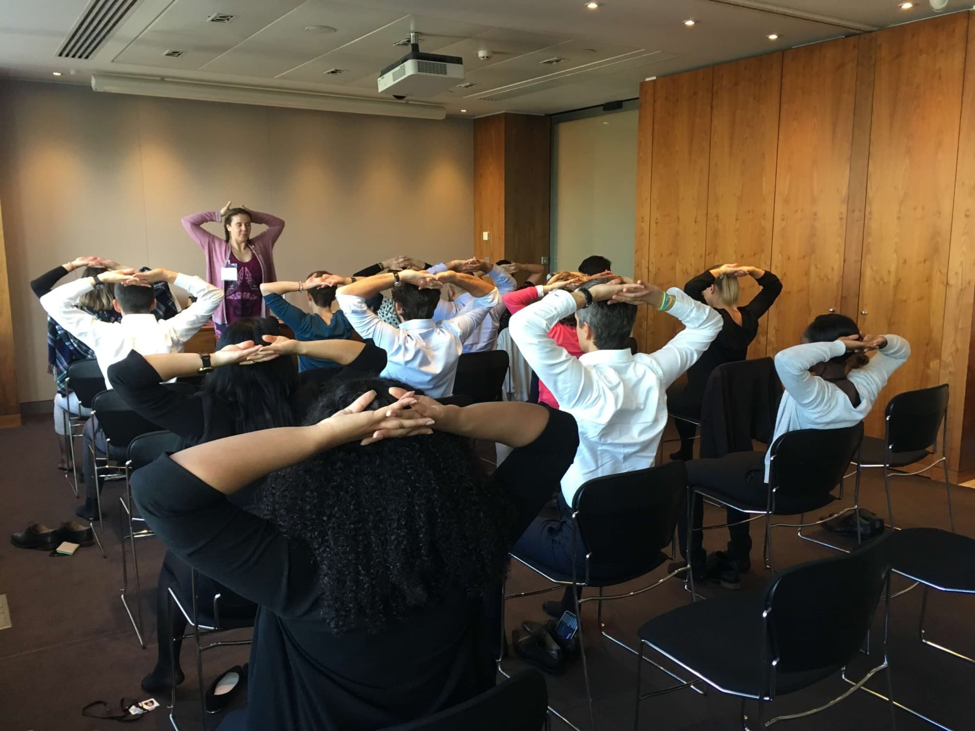 An instructor leading a seated chair yoga stretch for a group of employees in a meeting room